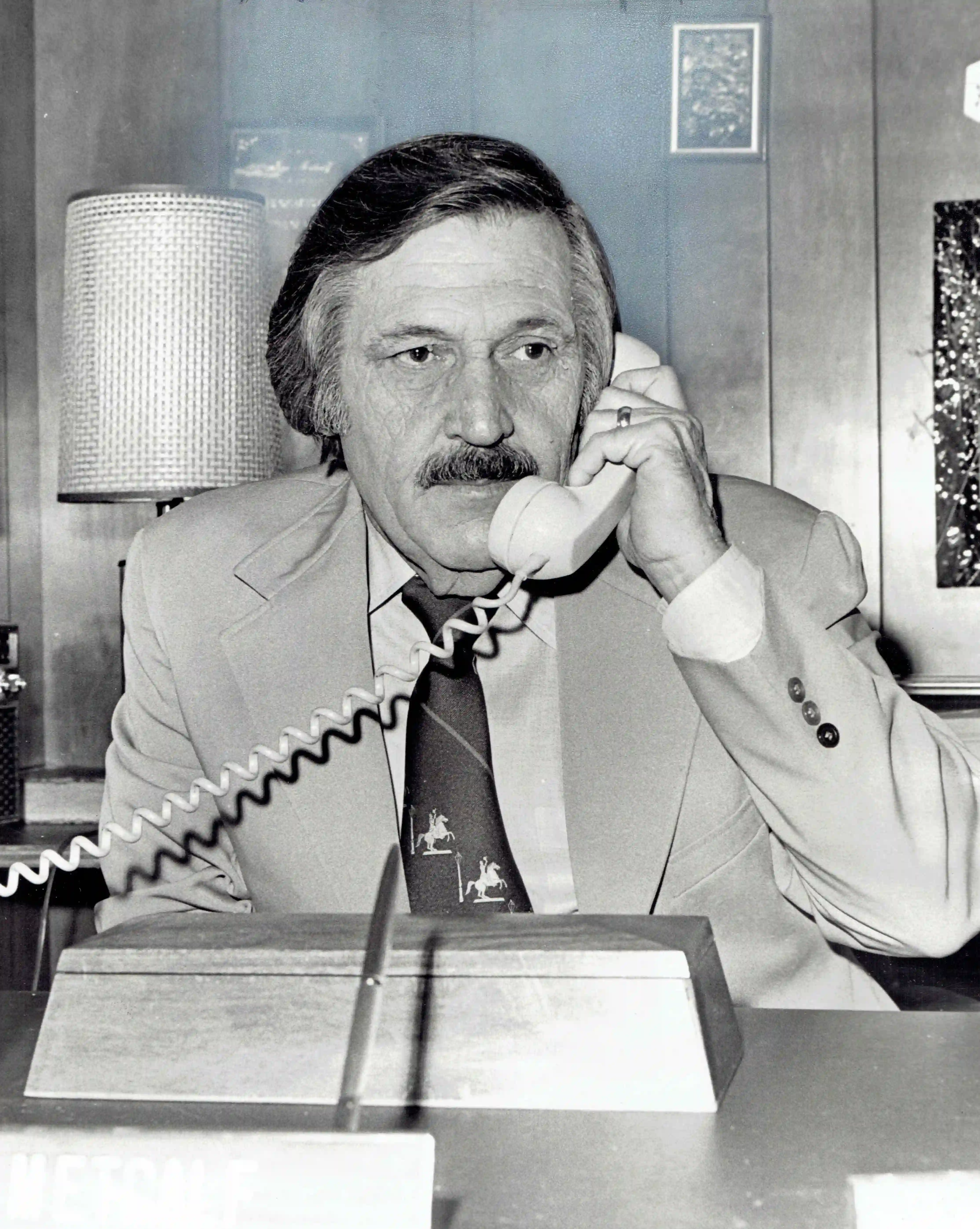Black and white portrait of Jim Metcalf from April 1975, as he takes a call at his desk, with a white phone receiver to his left ear with the coiled phone cable extending to his right, beyond the edge of the frame. His dark, greying hair is swept back from his face, which smiles only with his eyes, while his broad moustache sweeps the edge of the phone receiver. A dark tie boldly splits his light colored suit, which sports the broad lapel of the era. An unmarked wood box of writing accoutrements, with sloping trapezoidal sides sits in front of him, as he focuses his gaze to his left, off into the distance.
