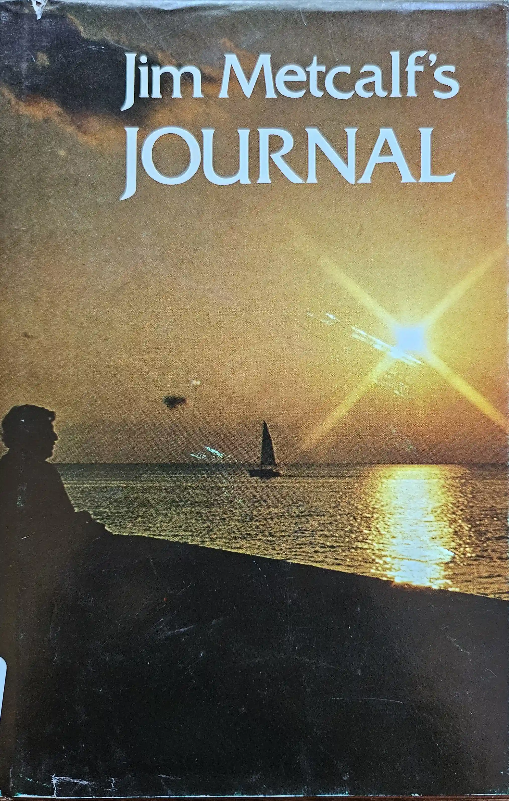 The front jacket cover of Jim's first book, 'Jim Metcalf's Journal' as captured in a close cropped photo. The front cover shows a warmly lit sunset from the south shore of Lake Ponchartrain in New Orleans. The shoreline, along with a lone figure on the shore and a sailboat in the distance, is darkened by the vivid exposure of the sun, leaving only a suggestion of the foreground. The title is featured at the top, in a font with very slight serifs and close kerning. The word 'JOURNAL' is in all-caps.
